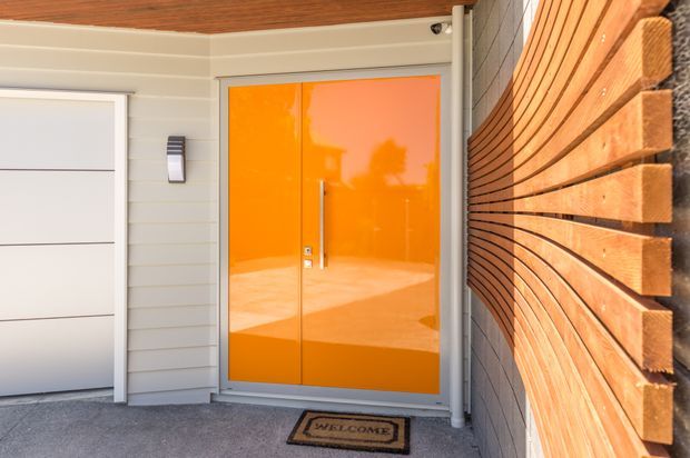 Contemporary residential entryway viewed from outside, featuring a vivid glossy orange double front door with slim vertical handles, pale grey horizontal cladding, a white sectional garage door, and a distinctive curved wall of warm timber slats. The covered porch has a grey concrete floor, wood-lined soffit, wall light, and welcome mat.