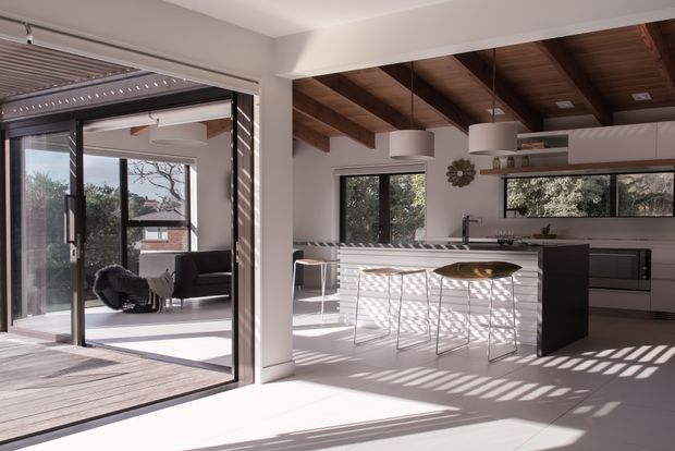 Bright, open-plan contemporary kitchen and living area with strong indoor-outdoor connection. The kitchen features a large white island with bar seating, dark integrated cabinetry, pendant lights, and a long horizontal window. Exposed timber ceiling beams and timber decking add warmth, while full-height black-framed sliding glazing opens onto an outdoor deck and a secondary lounge area. Strong sunlight creates dramatic linear shadows across the pale interior floor.