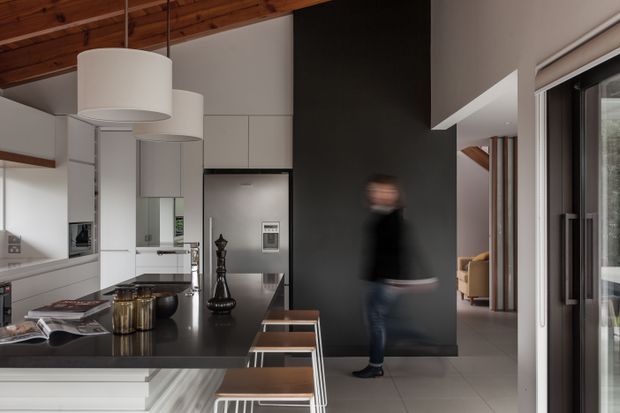Contemporary open-plan kitchen with a long dark island, white floor-to-ceiling cabinetry, integrated appliances, pendant lights, and a tall charcoal feature wall. A timber-lined sloping ceiling adds warmth, while a glazed opening connects the kitchen to an adjacent living area or hallway.