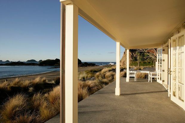 A covered coastal veranda or terrace overlooking a sandy beach and calm water. The long outdoor space has simple square columns, a flat ceiling, pale cream walls and French doors, with a white outdoor dining table and chairs near the rocky shoreline. Natural tussock planting surrounds the terrace, creating a relaxed seaside setting.