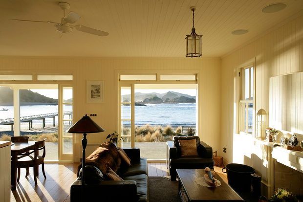 A warm, sunlit coastal living room and dining area with wide windows and glazed doors overlooking a beach, water, and rocky headlands. The room features a dark leather sofa and armchairs, timber flooring, light vertical wall panelling, a fireplace or built-in hearth with mantel shelving, a central coffee table, dining furniture, a ceiling fan, and a hanging lantern-style light. The interior has a relaxed traditional coastal character with strong natural daylight and ocean views.