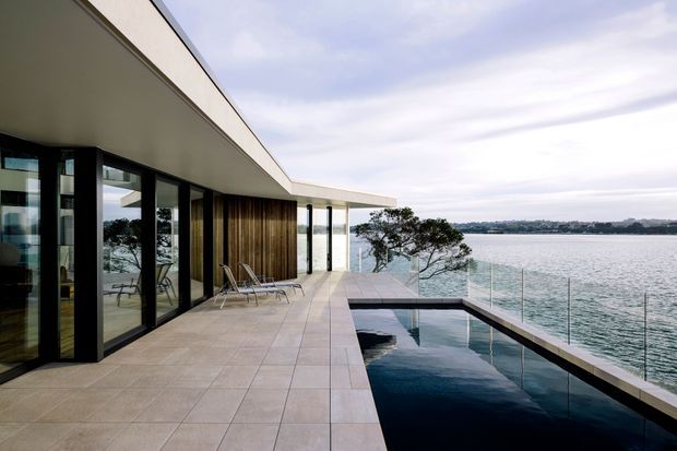 Contemporary waterfront residence with a broad tiled terrace, full-height dark-framed glazing, timber-clad wall panels, and a narrow infinity-edge swimming pool overlooking the water. The low, sweeping roof and transparent balustrade create a strong indoor-outdoor connection.