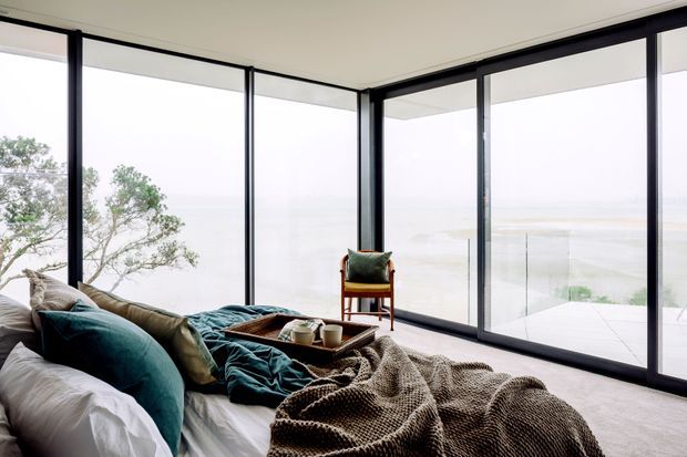 A serene bedroom corner with a bed layered in white linens, teal cushions, and a textured brown throw. Floor-to-ceiling black-framed glazing wraps around the room, opening to a bright coastal or waterfront outlook and a small balcony or terrace. A compact timber chair and tray table add warmth to the otherwise light, minimal space.
