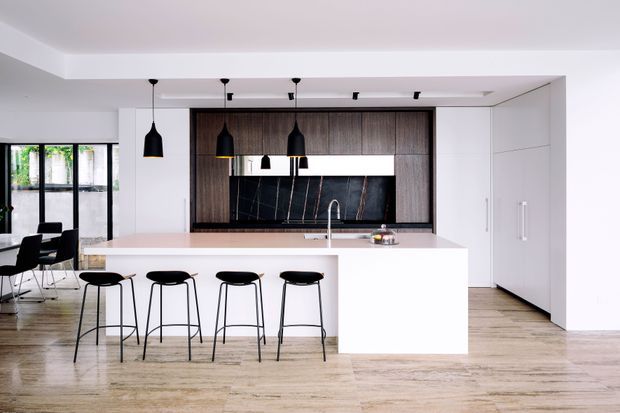 Bright contemporary open-plan kitchen with a large white island and four black bar stools, dark timber cabinetry, a black marble-look splashback, integrated white cabinetry, pendant lighting, and pale timber flooring. A dining area and glazed doors are visible to the left.