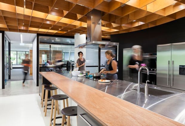 A spacious contemporary kitchen with a long timber island incorporating seating, a large stainless-steel benchtop and sink, dark cabinetry, integrated stainless-steel appliances, and a dramatic geometric timber coffered ceiling. Glazed openings connect the kitchen to adjacent bright spaces, while motion-blurred people suggest an active communal setting.