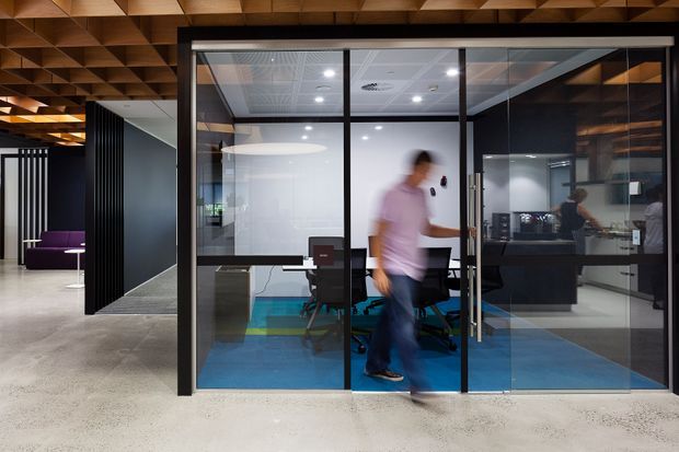 Contemporary commercial office interior viewed from a circulation area, with a glass-enclosed meeting or work room, black metal-framed glazing, blue carpet, dark furniture, and a distinctive exposed timber coffered ceiling. Open-plan office spaces and furnishings are visible beyond the glazed enclosure.