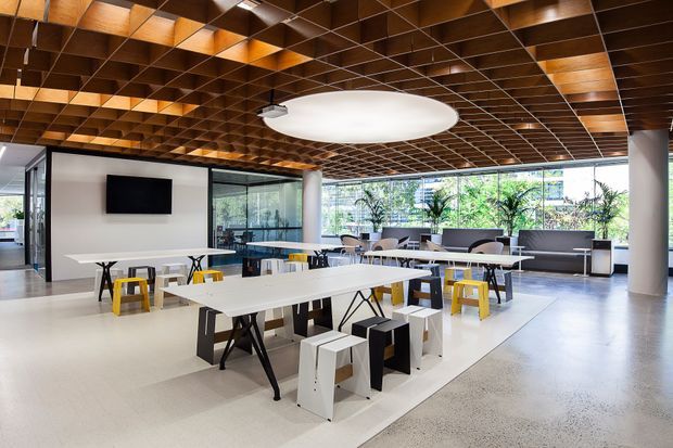 Open-plan contemporary commercial interior with communal dining or breakout tables, stools, a wall-mounted screen, extensive glazing, and indoor planting. The space features a distinctive coffered timber ceiling grid with integrated lighting, pale polished concrete or terrazzo-like flooring, white tables, and cylindrical structural columns.