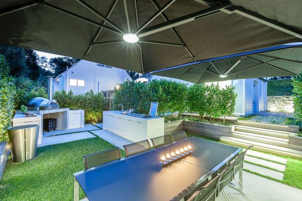 A landscaped outdoor entertaining area with a large dining table beneath overlapping shade umbrellas, an outdoor kitchen island with sink and grill, manicured hedging, stepped paving, and a modern light-coloured building in the background. The space is illuminated by integrated umbrella lighting and table candles, creating a polished evening entertaining setting.