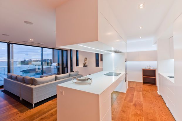 Bright open-plan contemporary kitchen and living area with extensive white built-in cabinetry, a large central island with integrated cooktop, pale benchtops, warm timber flooring, and a grey sectional sofa. Floor-to-ceiling glazing overlooks a waterfront setting, while recessed ceiling lights and clean-lined joinery create a refined minimalist interior.