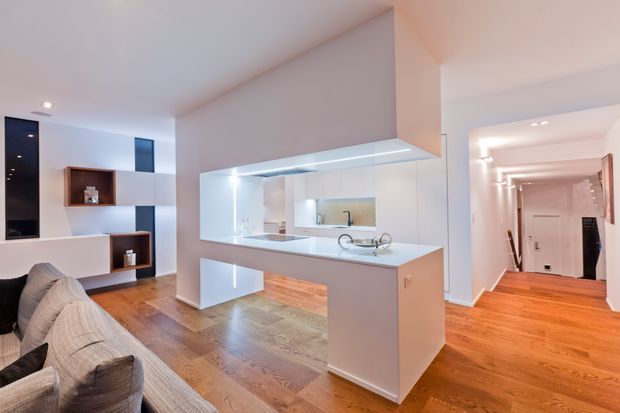 Bright open-plan contemporary interior featuring a compact white kitchen with a peninsula island, integrated lighting, pale stone surfaces and timber flooring. The kitchen opens to a living area with built-in shelving and a sofa, while a long illuminated hallway extends to the right.