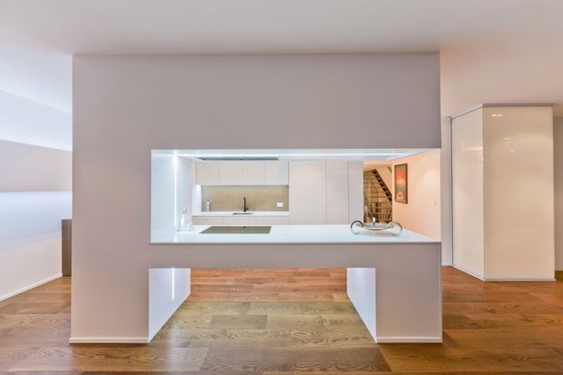 Minimalist open-plan interior featuring a white kitchen with a long island and integrated cabinetry, viewed through a large rectangular opening in a partition wall. Warm timber flooring, clean white surfaces, recessed lighting, and a staircase visible in the background create a refined contemporary feel.