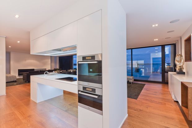 Open-plan contemporary interior showing a white kitchen with a large island, integrated cabinetry and stacked ovens, opening to a living area and a glazed waterfront-facing space. Warm timber flooring, white joinery, recessed lighting and dark furnishings create a refined, bright interior.