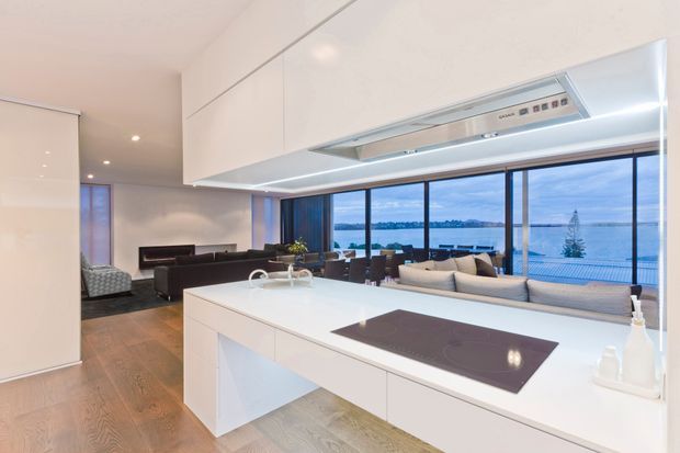 Bright open-plan kitchen with streamlined white handleless cabinetry, a large white island with integrated induction cooktop, and timber flooring. The kitchen opens directly to a contemporary living area and extensive floor-to-ceiling glazing, with a waterfront view visible beyond the balcony. The overall design is clean, modern, and minimal with cool blue exterior views and warm timber underfoot.