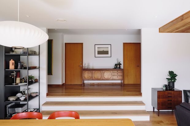Bright mid-century-inspired open-plan interior showing a dining area in the foreground, with a raised living or entry zone beyond. The space features warm timber flooring and doors, a sculptural white pendant light, open black shelving, a vintage-style timber sideboard, framed artwork, indoor plants, and a dark sofa at right. Broad stepped platforms create a clear level change between the dining and rear area.