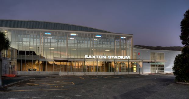 Exterior dusk view of Saxton Stadium, a large contemporary sports facility with a glazed multi-storey entrance facade, illuminated interior, prominent signage, and a ribbed metal-clad roof and side volume. The forecourt and parking area occupy the foreground.
