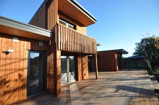 Exterior view of a contemporary timber-clad residential building with a projecting upper level, covered balcony enclosed by vertical timber slats, large glazed doors, and a broad timber deck or patio. The low-pitched roof forms and warm wood facade are clearly visible under bright daylight.