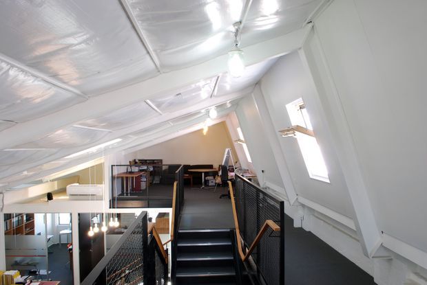 Bright loft-style interior with a central staircase leading to a mezzanine workspace. The space has white sloping ceilings, exposed structural framing, dark flooring, black mesh balustrades, timber handrails, and an industrial office fit-out with desks and shelving.