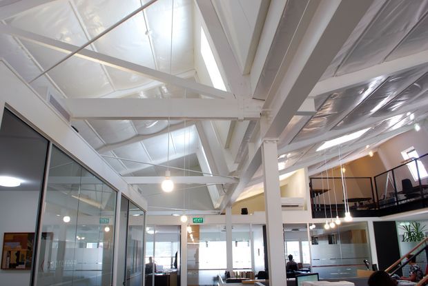 Bright contemporary commercial interior with an open-plan office layout, exposed white-painted structural beams and columns, a high industrial-style ceiling with visible reflective insulation, clerestory glazing, glass-partitioned rooms, pendant lighting, and a mezzanine level with black metal balustrades.