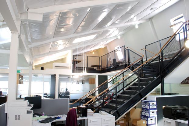 Bright open-plan commercial office interior with workstations, printers and storage in the foreground, a prominent black metal staircase with timber handrails, and a mezzanine level with additional desks. The space has exposed white-painted structural elements, abundant glazing, suspended pendant lights, and a partially exposed reflective ceiling insulation system.