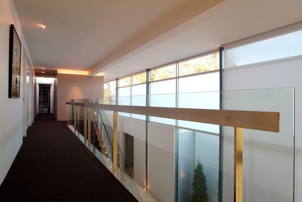 Interior view of a modern upper-level hallway or landing overlooking a double-height void. A dark carpeted corridor runs beside a glass balustrade with a timber handrail, while expansive clerestory glazing brings daylight into the space. Warm recessed and concealed lighting accents the white walls and ceiling, with stairs and a lower-level area visible below.