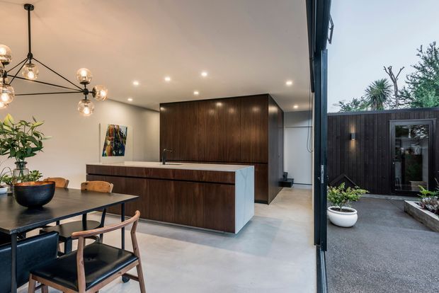 A contemporary open-plan kitchen and dining area opening directly onto a compact landscaped courtyard. The kitchen features dark timber cabinetry, a pale stone island with integrated sink, polished concrete flooring, and a sculptural multi-globe pendant above the dining table. Full-height glazing and an open glazed door connect the interior to a dark-clad courtyard with built-in garden beds and potted plants.