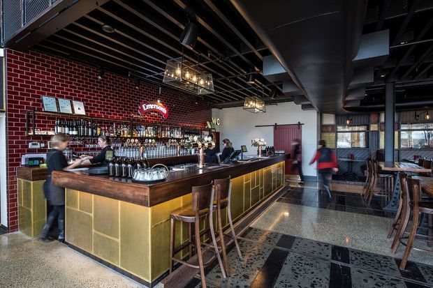 Interior of a stylish bar or restaurant with a prominent L-shaped timber bar, brass-toned panelled front, exposed black ceiling services, red brick feature wall, beer taps, pendant lighting, and scattered dining tables and chairs. The space has an industrial-inspired fit-out with warm hospitality detailing.
