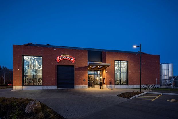 Exterior dusk photograph of a contemporary single-storey red-brick commercial building, featuring large glazed display windows, a recessed illuminated entrance canopy, a prominent brick facade, and a branded sign. The building sits beside a paved parking and access area with industrial tanks visible in the background.