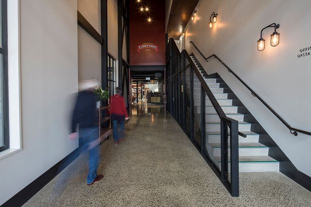 Interior view of a contemporary commercial passageway with polished terrazzo flooring, white walls, black metal-framed glazing, and a straight stair with black mesh balustrade. Industrial-style wall and pendant lighting line the corridor, with blurred people moving through the space and signage visible in the distance.