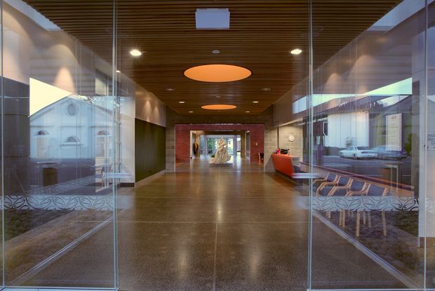 A contemporary commercial lobby or entry corridor viewed through open glass doors. The space has polished dark flooring, transparent glazed walls, a warm timber slatted ceiling with recessed and circular pendant lighting, dark wall panels, and a vivid red reception desk at the far end. Reflections and printed graphics are visible on the glass partitions, with seating along the sides.