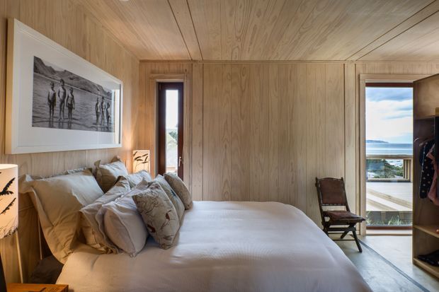 A compact bedroom clad throughout in warm natural timber, with a built-in timber-panelled wardrobe wall, a low bed with layered neutral bedding, a framed black-and-white artwork, and narrow vertical windows. Full-height glazing at the right opens views toward the ocean and coastal landscape. The room has a calm, warm, minimalist cabin-like character.