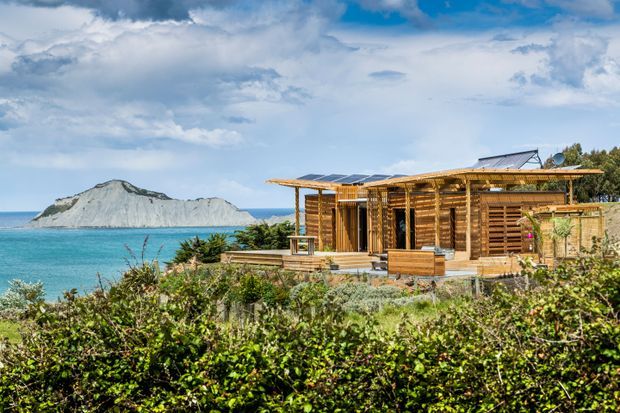 A low-slung timber-clad coastal dwelling set beside vivid turquoise water, with extensive timber decking, pergola-like roof framing, solar panels, and louvred timber screens. A distinctive pale rocky island or headland is visible offshore, while dense coastal planting fills the foreground. The architecture has a strongly site-responsive, relaxed and sustainable coastal character.