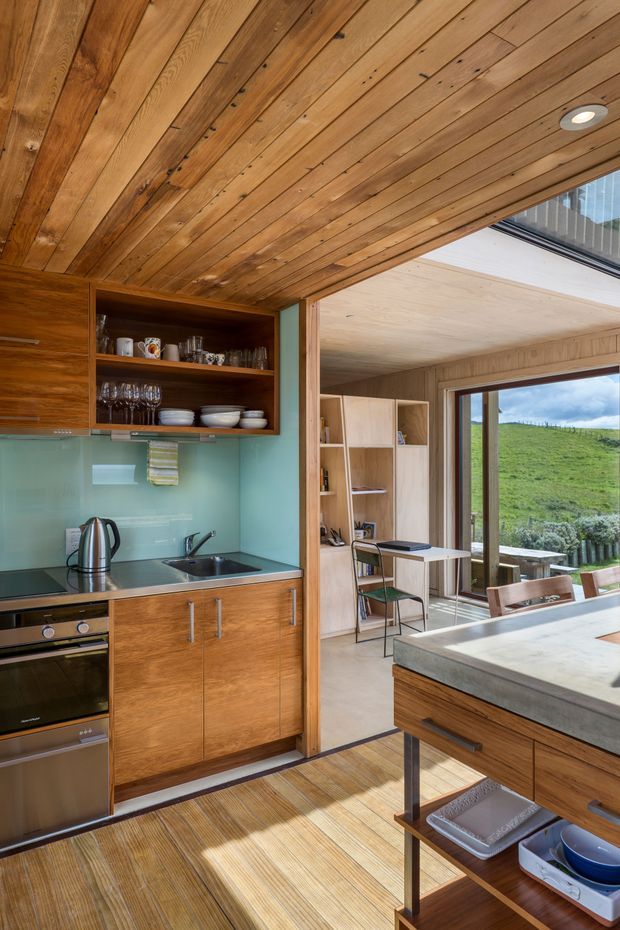 A warm, compact contemporary kitchen with extensive timber cabinetry and timber-lined ceiling. The kitchen includes a dark stone benchtop, stainless-steel sink and tap, integrated oven, open shelving with crockery and glassware, and a pale aqua glass splashback. A timber island or peninsula occupies the foreground. Beyond the kitchen is a light timber-lined dining or living area with built-in shelving, clerestory-style glazing, and views toward a green hillside.
