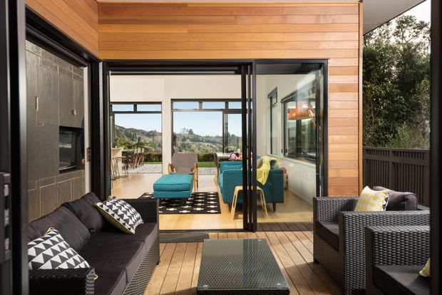 A contemporary indoor-outdoor living area with a timber-lined ceiling and walls, dark-framed sliding glass doors, and a timber deck. Outdoor wicker seating surrounds a low coffee table in the foreground, while a bright living room with teal seating and patterned rugs is visible beyond the glazing. The space opens toward a landscaped hillside view.