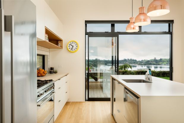 Bright contemporary kitchen with white cabinetry, a large central island, stainless-steel appliances, timber flooring, and black-framed glazing overlooking a waterfront landscape. Three copper pendant lights hang above the island, while open shelving and a yellow wall clock add visual accents.