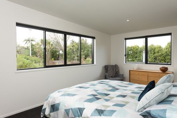 Bright contemporary bedroom with large black-framed windows overlooking greenery, a low timber dresser, upholstered armchair, and a bed dressed in a geometric blue, grey, and white quilt. The room has simple white walls, a plain ceiling with recessed lighting, and dark carpet flooring.