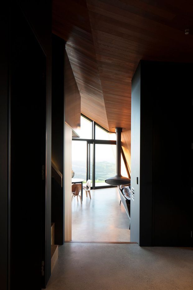A dark entry view looking into a minimalist open-plan living and dining space. The room features a warm timber-lined sloping ceiling, extensive black-framed floor-to-ceiling glazing, polished concrete flooring, a suspended black fireplace, and a dramatic elevated landscape view beyond. The interior has strong contrast between the dark entry joinery and the bright, airy living area.