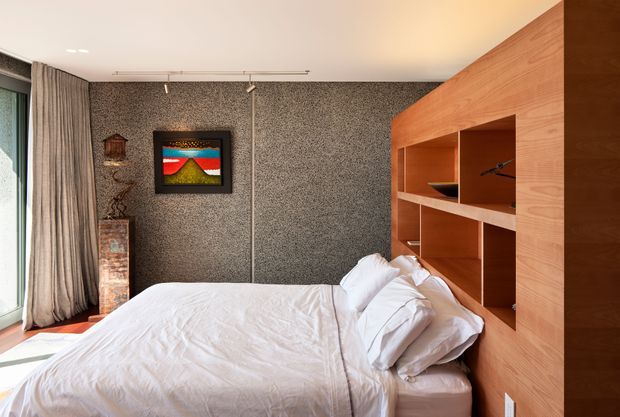 A compact contemporary bedroom with a white bed positioned against a full-height warm timber built-in headboard and open shelving. The room features a dark textured feature wall, pale curtains beside a large window, recessed display niches, track lighting, and restrained decorative artwork. The overall composition is warm, refined, and slightly moody.
