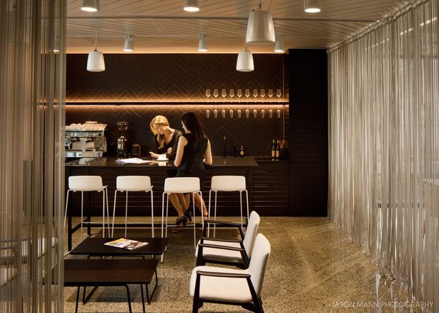 A sophisticated commercial bar and lounge interior with a dark timber bar, illuminated shelving, hanging pendant lights, white bar stools, and a small seating area. Vertical sheer drapery screens frame the space, while a patterned timber ceiling and textured carpet add visual interest. Two people are present at the bar.