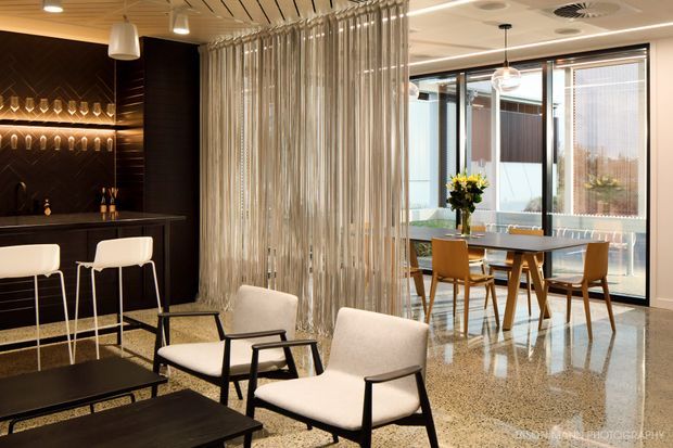 Refined open-plan hospitality or commercial interior with a dark built-in bar, illuminated bottle display, sculptural pendant lights, terrazzo flooring, and a dining area beside full-height glazing. A curtain of vertical metallic rods separates the lounge seating from the dining space, creating a luxurious contemporary atmosphere.