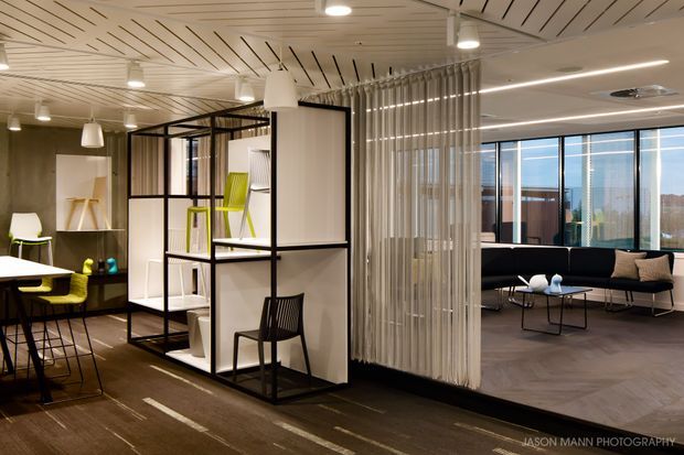 A contemporary open-plan commercial interior with a collaborative dining or breakout area on the left and a lounge area on the right. Black-framed modular shelving and translucent vertical curtains divide the spaces, while large glazed windows, pendant lights, linear ceiling lighting, and acoustic slatted ceiling panels create a bright, refined workplace environment.