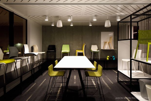 A contemporary commercial-style dining and meeting space with a large white rectangular table surrounded by lime-green and black chairs. The room features dark carpet flooring, a textured charcoal feature wall, black-framed glass partitions, open black shelving, a white slatted ceiling, and multiple pendant and track lights. Additional chairs and display furniture line the perimeter, creating a showroom or hospitality-oriented setting.