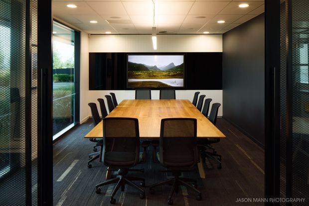 A contemporary boardroom with a long timber conference table surrounded by black office chairs. The room has dark feature walls and joinery, floor-to-ceiling glazing on one side, recessed ceiling lights, and a wall-mounted display screen showing a mountain landscape.