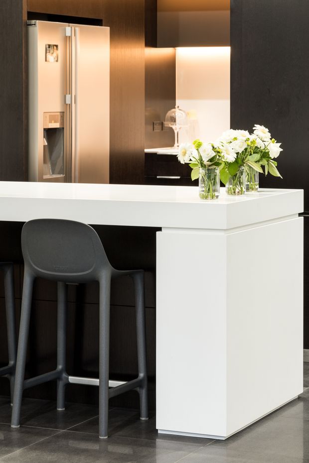 Contemporary kitchen interior focused on a white island or breakfast bar with a thick waterfall-style end panel and dark integrated cabinetry. Two dark stools sit beneath the overhang, while a stainless-steel refrigerator, illuminated splashback or work area, and glass flower arrangements are visible behind. The space has polished dark flooring, a restrained black-and-white palette, and warm concealed lighting.