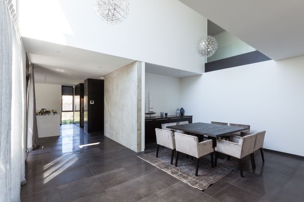A bright, spacious contemporary dining area with a dark tiled floor, large rectangular dining table, upholstered dining chairs, and a textured rug. The room opens into a hallway and kitchen or living area, with dramatic high ceilings, a partial upper-level void, white walls, dark built-in joinery, and spherical wire pendant lights.