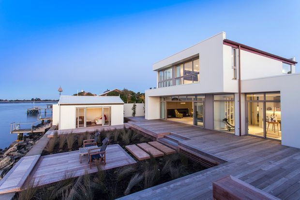 A contemporary multi-level waterfront residence with crisp white rendered walls, extensive floor-to-ceiling glazing, and a low-pitched roof. Broad timber decking wraps around the home and extends over landscaped beds, with a small separate pavilion beside the water. The scene includes outdoor seating, a calm waterfront setting, and a refined indoor-outdoor connection.