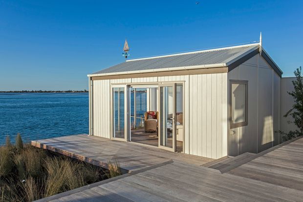 A compact waterfront pavilion or small dwelling with pale vertical cladding, a steep gabled metal roof, and large glazed sliding doors opening onto a broad timber deck. The building sits directly beside calm blue water, with sparse coastal planting around the deck. A small furnished interior is visible through the glazing.