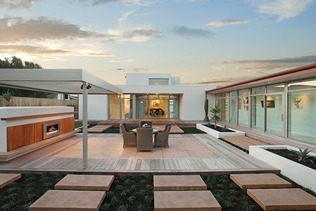 Contemporary courtyard-style outdoor living area at dusk, framed by low white modern buildings with extensive glazing. The landscaped terrace includes tiled paving, timber stepping-stone paths, built-in raised garden beds, a freestanding dining setting, and a covered outdoor fireplace or barbecue zone.