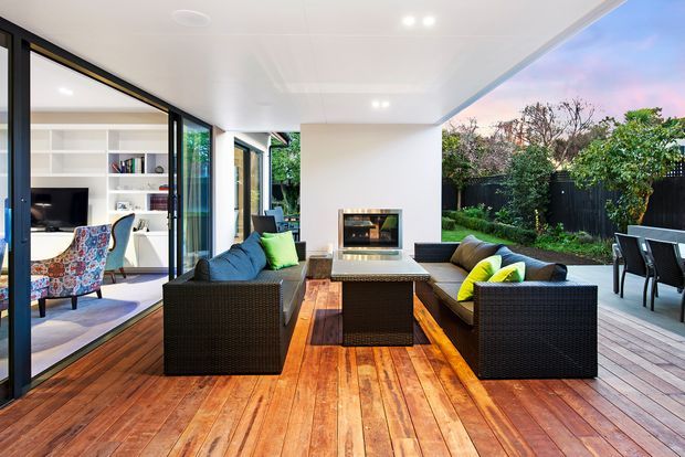 A bright contemporary indoor-outdoor living area opening onto a timber deck and garden. The covered space features a white ceiling, open-sided glazing, a built-in fireplace, dark woven outdoor-style sofas and armchairs, a central coffee table, and colourful cushions. An adjacent living room with shelving and a television is visible through the glazed opening.