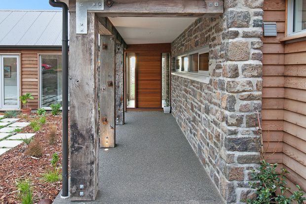 Covered residential entry walkway with a textured grey floor, heavy timber posts and beams, rough stone masonry walls, horizontal timber cladding, narrow clerestory-style windows, and a timber front door. A landscaped courtyard and adjoining timber-clad wing are visible to the left.