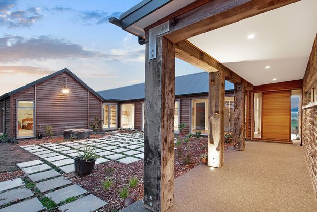 A rustic contemporary residential courtyard and covered entryway. The building features horizontal timber cladding, dark metal pitched roofs, substantial weathered timber posts and beams, a stone feature wall, warm timber doors, and a patterned concrete paver path through landscaped gravel. The covered corridor opens toward a scenic landscape beyond.